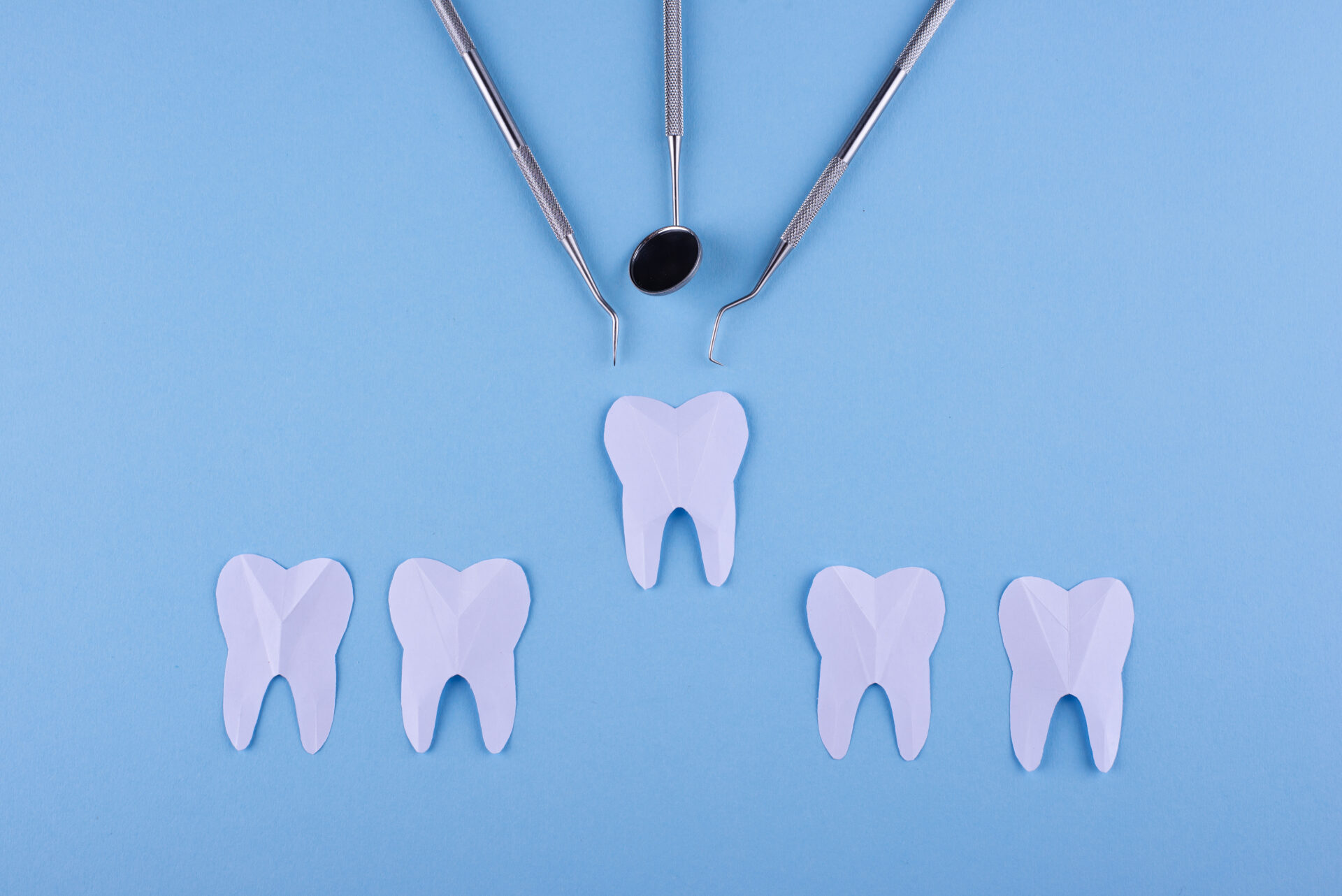 Paper cut-out teeth beneath three dental instruments on a blue background